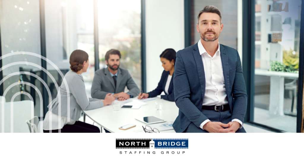 Employer in corporate attire leans on the edge of an office meeting table, smiling confidently