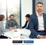 Employer in corporate attire leans on the edge of an office meeting table, smiling confidently