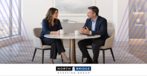 Two professionals smile as they talk over coffee at a round marble table in a bright office. North Bridge Staffing Group logo.